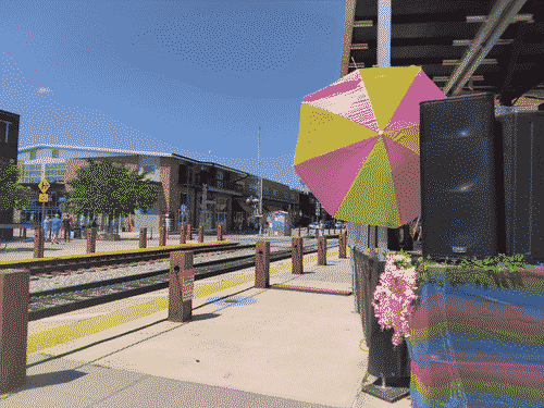 Colorful stage/AV setup for Sol Sunday with flowers, reflective mesh fabric, multicolored umbrella, etc, with the railyard district in the background.