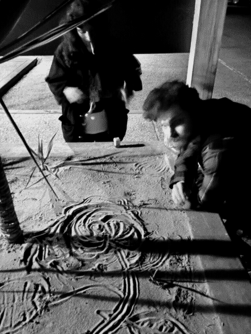 Brandon leans on a xeriscape planter where he and John (taking the photo) had collaborated on a sand drawing of a rose. Iris is behind the planter trying to light a joint