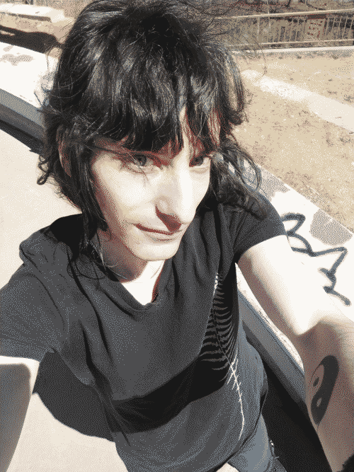 High angle photo of a dark haired, fair skinned transfemme enby with bangs, looking at the camera w/ green eyes. They are wearing a black shirt sleeved shirt with a gold fern leaf on the left side and black jeans. the top edge of a black inline skate is visible at the bottom of the frame. On the inside of their left arm is visible a black yin-yang tattoo. In the background is a C-ledge with graffiti on it typical of a street course at a skate park