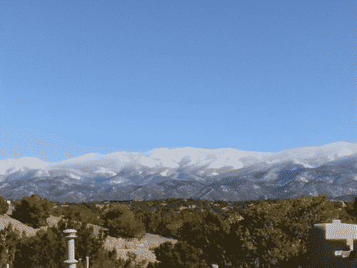 Snow-covered peaks east of Santa Fe against a cloudless blue sky, with the typical evergreen scrubland in the foreground