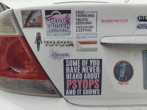 Left half of the backside of a white late-2000s Toyota camry, with various stickers. The most prominent reads 