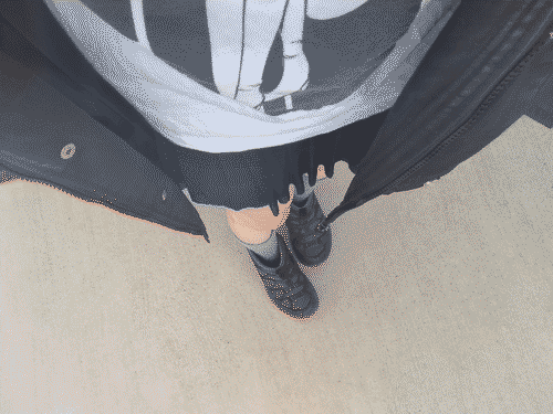 POV selfie of Iris looking down at themself - they are wearing a long black coat over a white graphic tee, black short mesh skirt, grey socks, and black shoes. Their thighs are creamy in the midday light against their dark clothes.