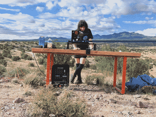 Iris at a freestanding wood-topped DJ table, leaning against it operating the keyboard, with a smirk on her face as she selects a track, her crossed legs visible beneath the table. She is wearing a black shirt, black shorts, black thighhigh socks, and black leather midtop boot, and her dark hair hangs unbound around her face. Below the table is also a PA system