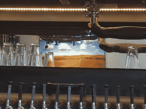 A mirror shows an impressonist desert or prairie landscape, framed above bypipes and a row of LED lights, below by beer glasses and beer taps which are made of railroad spikes