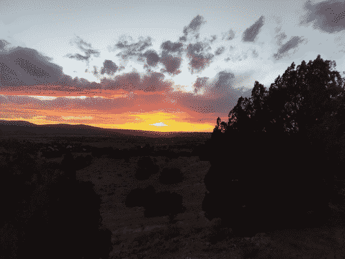 Brilliant orange sunset with pink clouds above, flanked on the left by faintly purple mountains and below by the rolling chapharelle hills of Cerillos