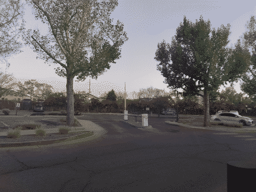 A road barrier for preventing unauthorized entry into a parking lot, lit up with red LEDs in the twilight and stuck in the vertical, open, position. The parking lot itself is mostly empty and two healthy deciduous trees flank the entrance to the parking lot.