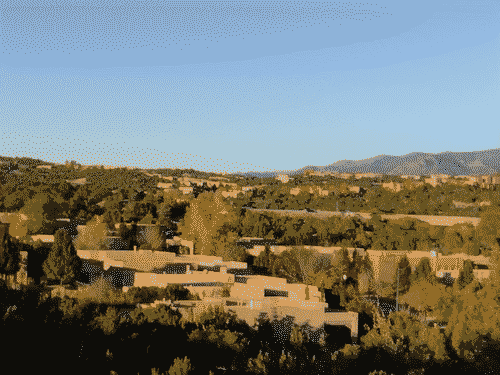 Arrays of typical upscale Santa Fe houses embedded in the pine and Juniper forest of northern Santa Fe, creating a terraced network of beige blocks in the bursting green of the trees. A duller green of the mountains rises sinusoidally along the right edge of the horizon, and the sky is clear blue.