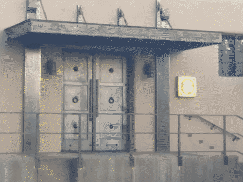 A cryptic facade with ornate, heavy steel double doors flanked by modern reflective lamps and heavy steel columns, and an awning which is also heavy steel and held in place by tension from cables attached from the awning at a 45-degree angle to the wall above it. A yellow and grey glyph (undecipherable in part due to the lighting) is embedded in the wall to the right of the gates, which wall is fake adobe.