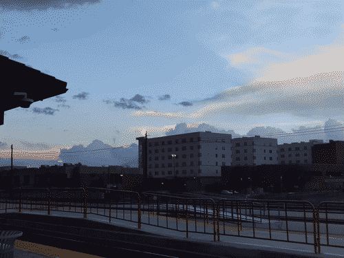 View across the downtown train platform in Albuquerque, with multiple tracks and platforms in the foreground along with a security camera on the corner of a weather-protected seating area. Brutalist architecture lies beyond, blocky and featureless except the grid of windows, with a hint of sunset in the low-hanging clouds to the left of the buildings.