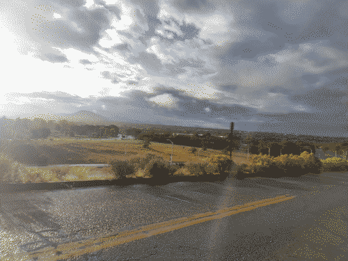 View atop Calle Mejia across Santa Fe after a night of rain. The moisture is palpable in the mist as well as the sheen on the surface of the road, and the sun peeks out of the dispersing rain clouds above the green mountains.