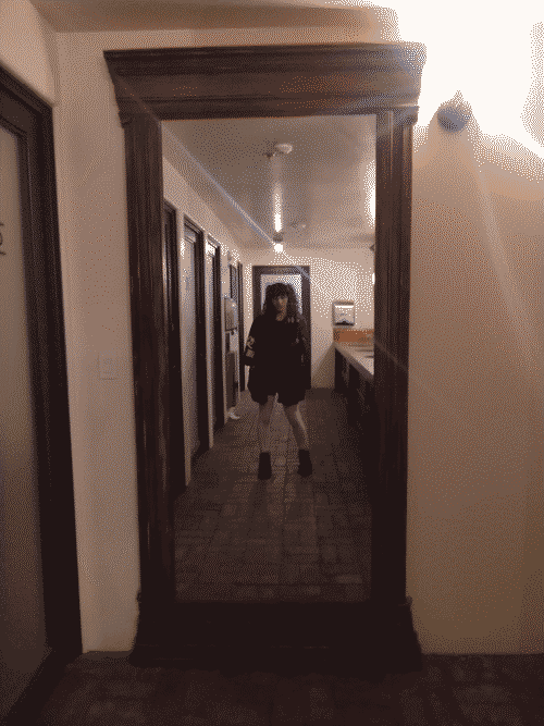 Iris taking a selfie in the full-length mirror in the large unisex bathroom of the lower level of Nuckolls Brewery. She is wearing her dark hair in twintails, her black coat with 3/4 sleeves, black shorts, white stockings which catch the light beautifully and accentuate the curves of her legs, and black midtop boots. She holds the phone next to her head in a fingerlessly-gloved hand, her other hand similarly clad, hanging at her side with kandi bracelets on the wrist. The floor is brick, and the mirror is framed in heavy ornate wood against the off-white wall. Visible in the reflection flanking Iris are the rows of frosted glass doors to the stalls on one side and the rows of sinks embedded in the counter on the other side. 