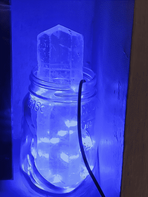a large selenite crystal is lit up blue inside of a mason jar by an LED strip that snakes around it inside the jar