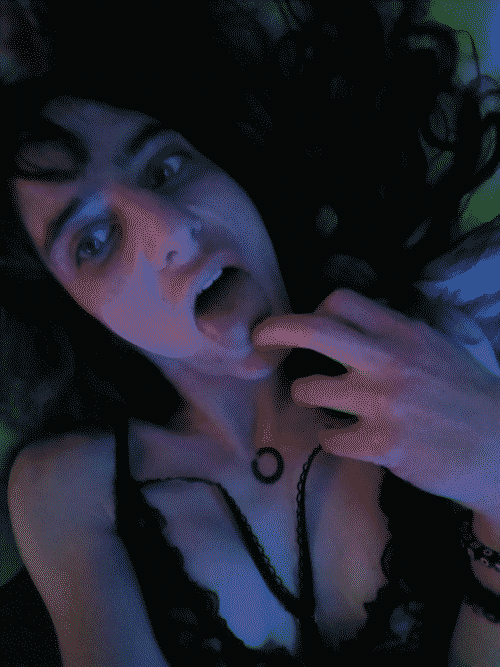 Bust portrait of Iris in lingere licking the tip of one of her fingers while she looks into the camera seductively. Her hair is splayed behind her on the bedspread and her breasts are pushed up by gravity since she is laying down. Her oroborous tattoo is visible in the crux of the Y of lacy fabric that comes together in her cleavage.