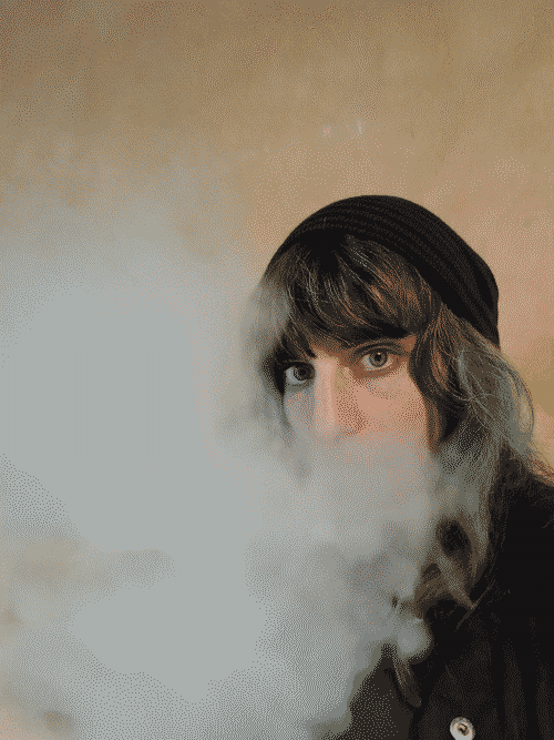 Iris in black jacket, striped black and purple beanie, and heavy mascara, breathing white vapor out in front of herface so that only her eyes are visible throug the cloud.