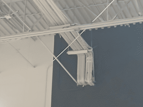 White corrugated metal cieling with cables and tresses running under it, and a large cluster of pipes creeping down the blue wall from the cieling where they meet, and then entering the wall.
