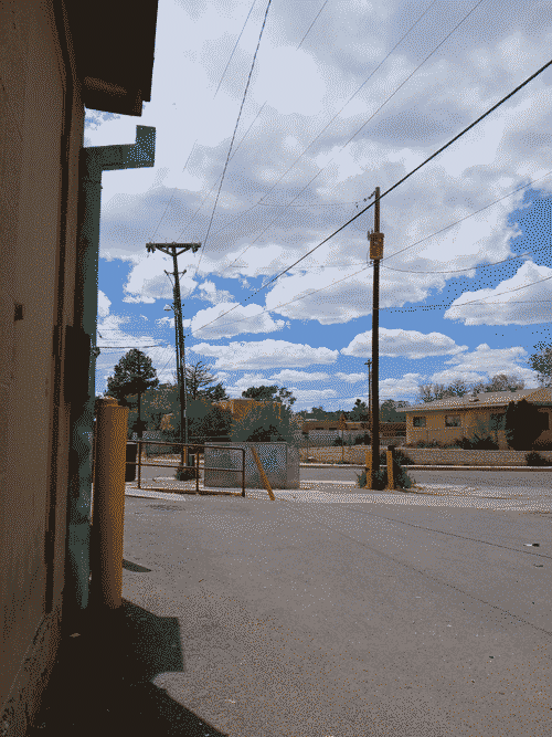power lines cross a cloudy sky from top-right, hugged by power poles as they veer left; a neighborhood with boxy Santa Fe houses and plenty of trees is visible across the recieving lot that dominates the foreground; a wall with a downward gutter attached and a yellow concrete pylon next to it runs along the left edge of the image
